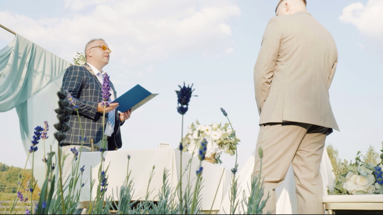 Plenerowa ceremonia ślubna: mężczyzna w okularach i kwiecistym wieńcu czyta z niebieskiej teczki, a za nim stoi pan młody w beżowym garniturze, w tle dekoracje i kwiaty.