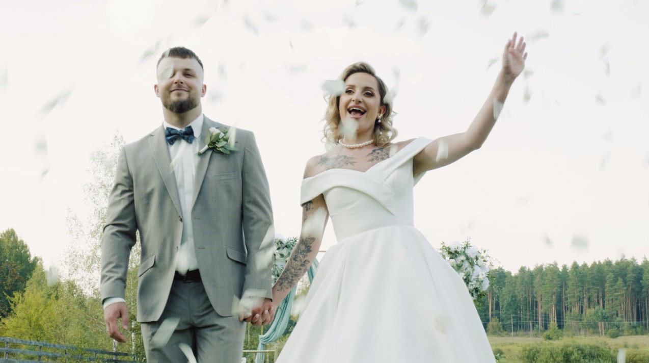 Para młoda, on w szarym garniturze z muchą, ona w białej sukni, trzymają się za ręce podczas ceremonii ślubnej, w powietrzu unoszą się płatki.