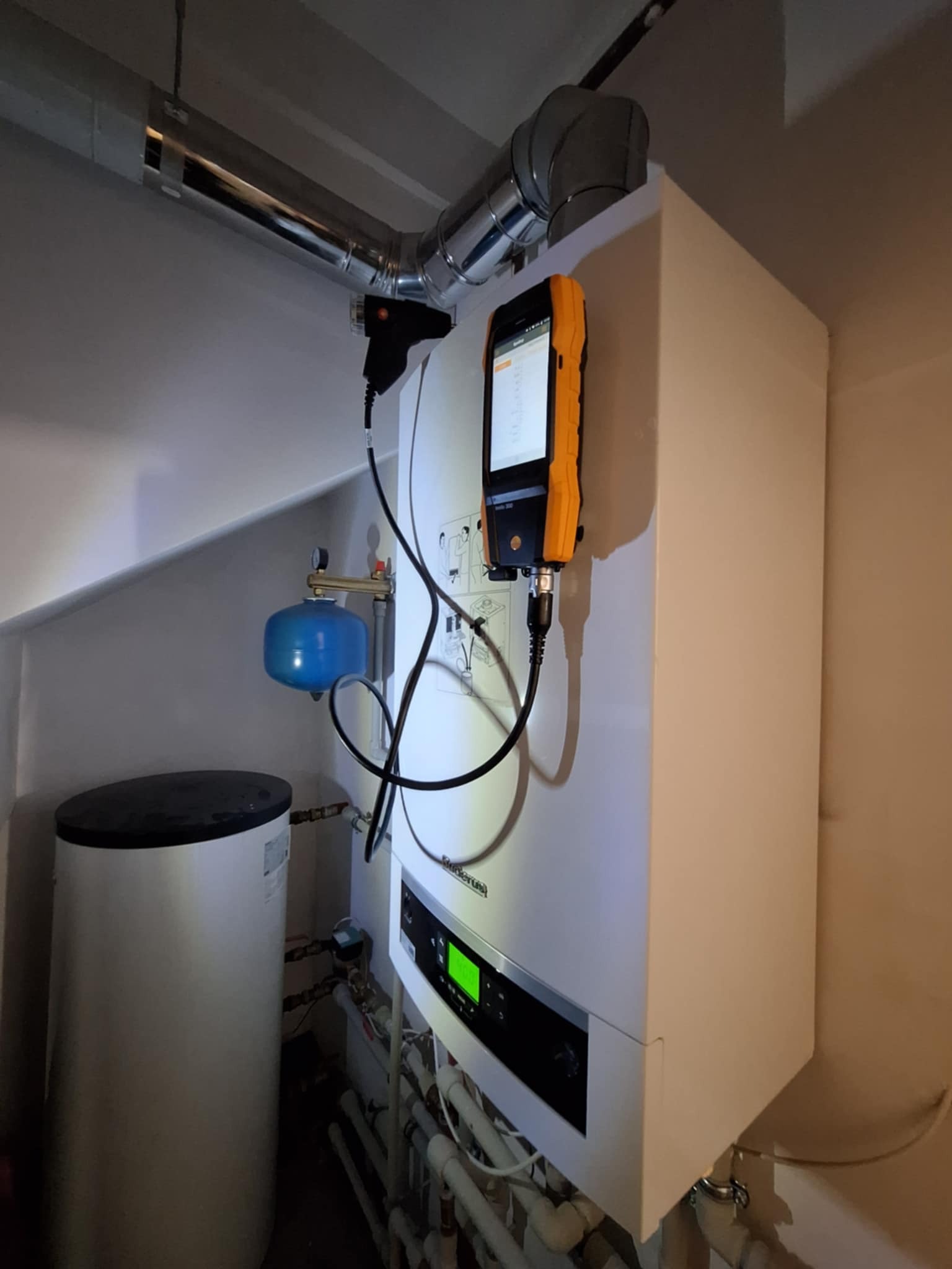 Biały piec grzewczy z podłączonym analizatorem spalin, widoczne rury i zbiornik, oświetlenie górne. Diagnostyka i serwis instalacji grzewczej.