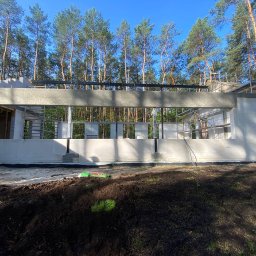 Dombud - Wznoszenie ścian domu z betonu komórkowego w otoczeniu drzew. Widoczne rusztowania i elementy konstrukcyjne. Etap budowy w lesie przy słonecznej pogodzie.