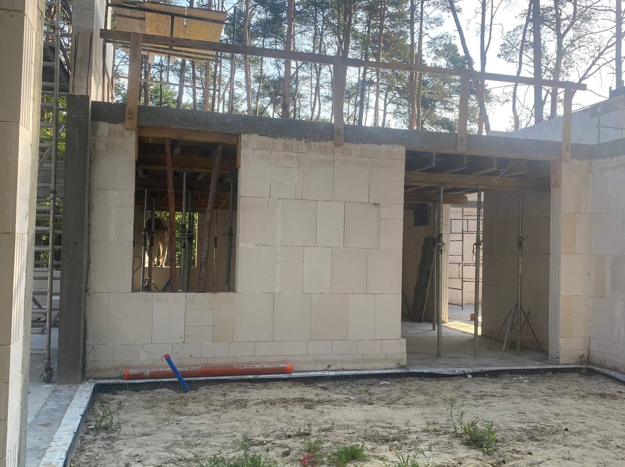 Surowy stan domu z betonu komórkowego w lesie, widoczne podpory okienne i drewniane szalunki stropu. Rury na piasku. Etap budowy w otoczeniu drzew.