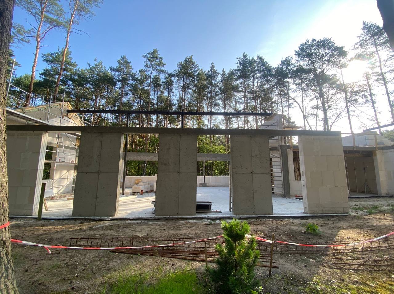 Budowa domu w lesie: surowe ściany z betonu komórkowego, widoczne zbrojenia i szalunki, etap wznoszenia konstrukcji. W tle wysokie drzewa.