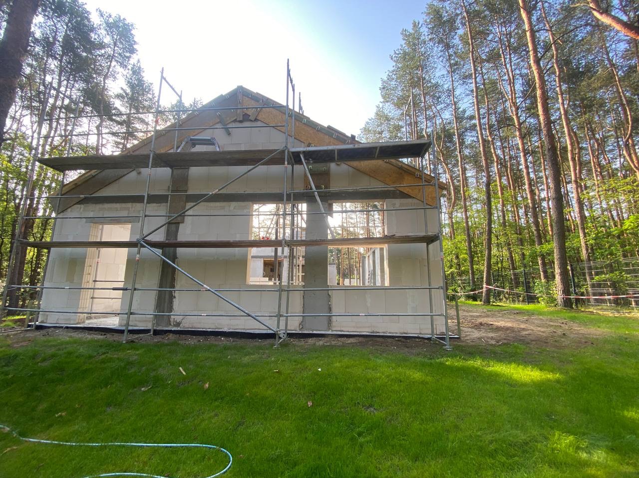 Budowa domu z białych bloczków, częściowo otynkowany, z drewnianą konstrukcją dachu, otoczony lasem i rusztowaniami. Widok z poziomu trawnika, w słoneczny dzień.