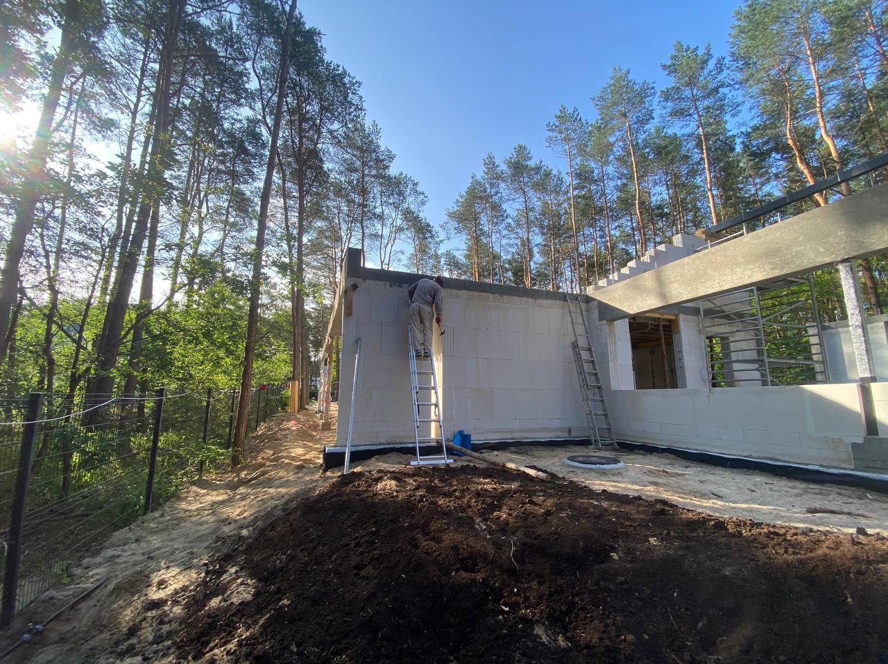 Budowa domu z betonu komórkowego w lesie. Mężczyzna na drabinie przy ścianie. Widoczne zbrojenia, szalunki i elementy konstrukcyjne. Słoneczny dzień.