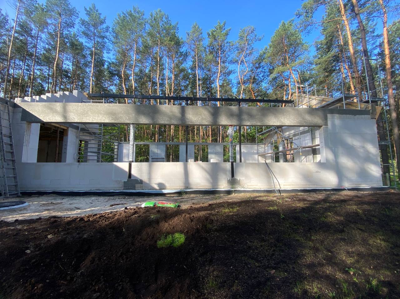 Wznoszenie ścian domu z betonu komórkowego w otoczeniu drzew. Widoczne rusztowania i elementy konstrukcyjne. Etap budowy w lesie przy słonecznej pogodzie.