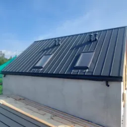 Dom w budowie z ciemnym, metalowym dachem z dwoma oknami dachowymi i wentylacją. Widoczne rusztowania i elementy konstrukcyjne.