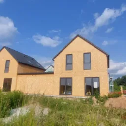 Dwa domy szkieletowe w stanie surowym zamkniętym, z czarnym dachem i oknami, na tle błękitnego nieba z chmurami. Na pierwszym planie trawa i materiały budowlane.