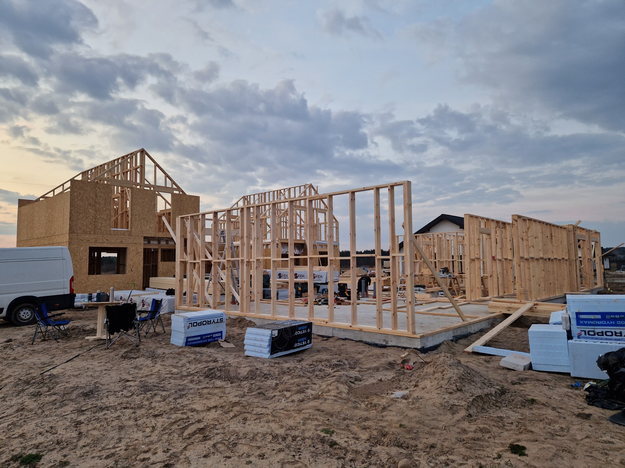 Budowa domu szkieletowego: drewniana konstrukcja w trakcie montażu, widoczne płyty OSB, styropian, narzędzia i pojazd dostawczy. Niebo z chmurami w tle.
