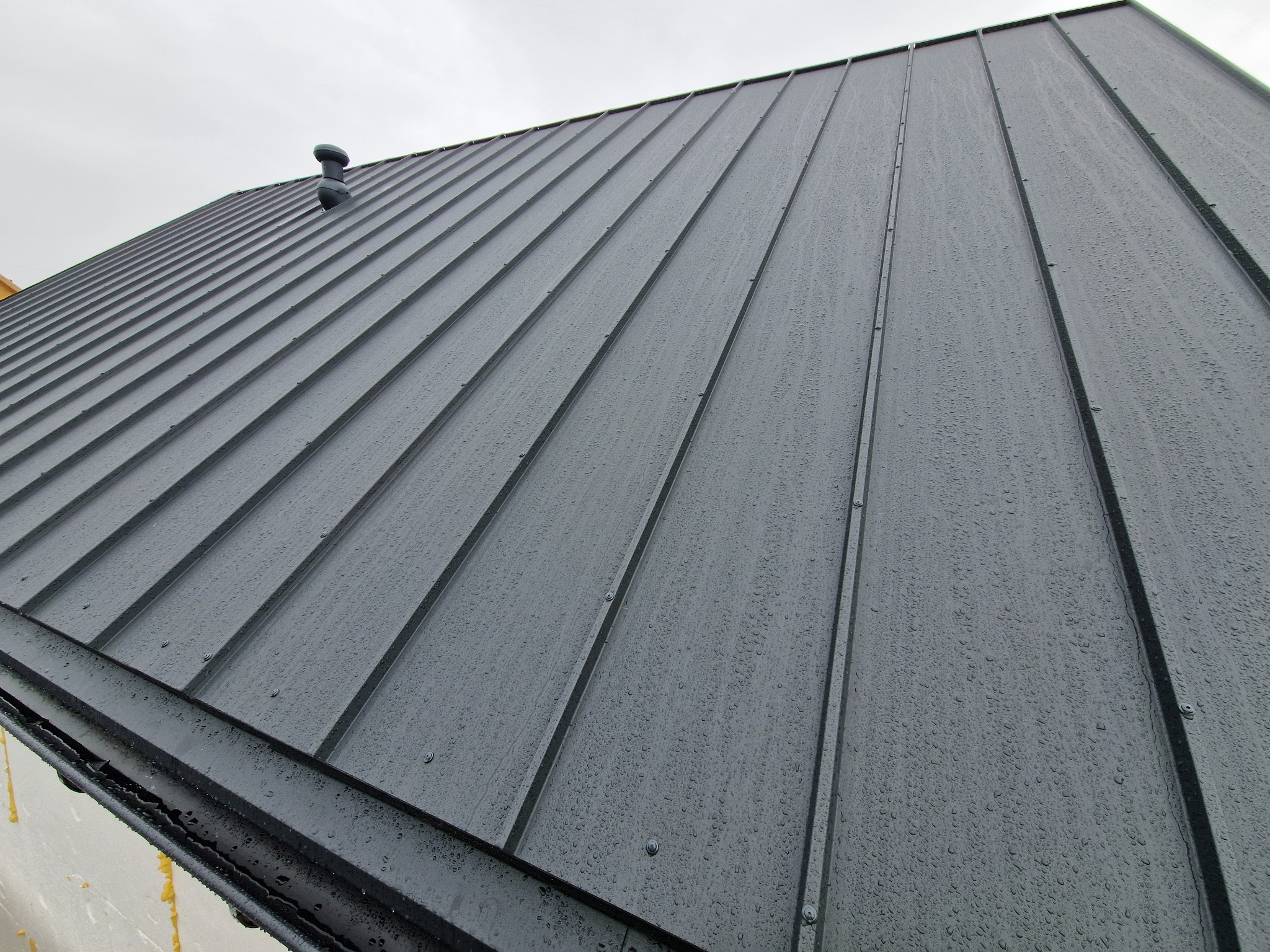 Szary, metalowy dach z widocznymi kroplami deszczu, ułożony pod kątem. W tle fragment nieba i komin wentylacyjny. Nowoczesne krycie dachu.