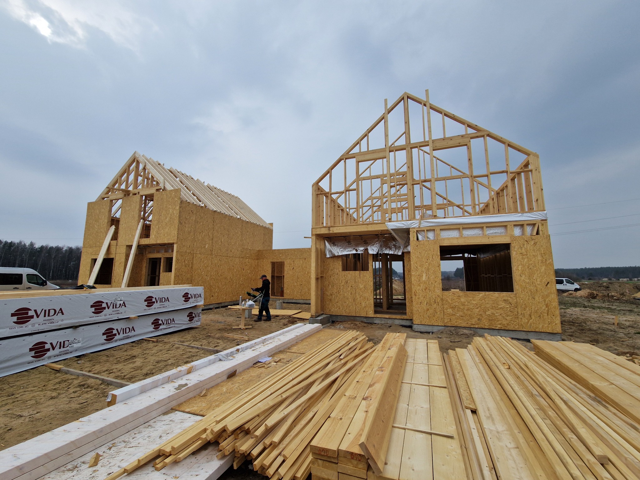 Budowa domu szkieletowego w trakcie: widoczne drewniane elementy konstrukcyjne, ściany z płyt OSB i materiały budowlane. Pracownik przy pracy.