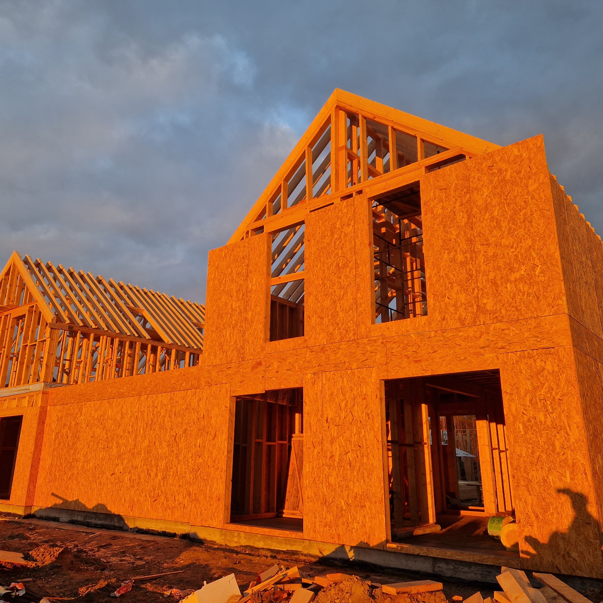 Wczesny etap budowy domu szkieletowego, widoczne drewniane elementy konstrukcyjne i poszycie ścian w ciepłym świetle zachodzącego słońca, na tle pochmurnego nieba.