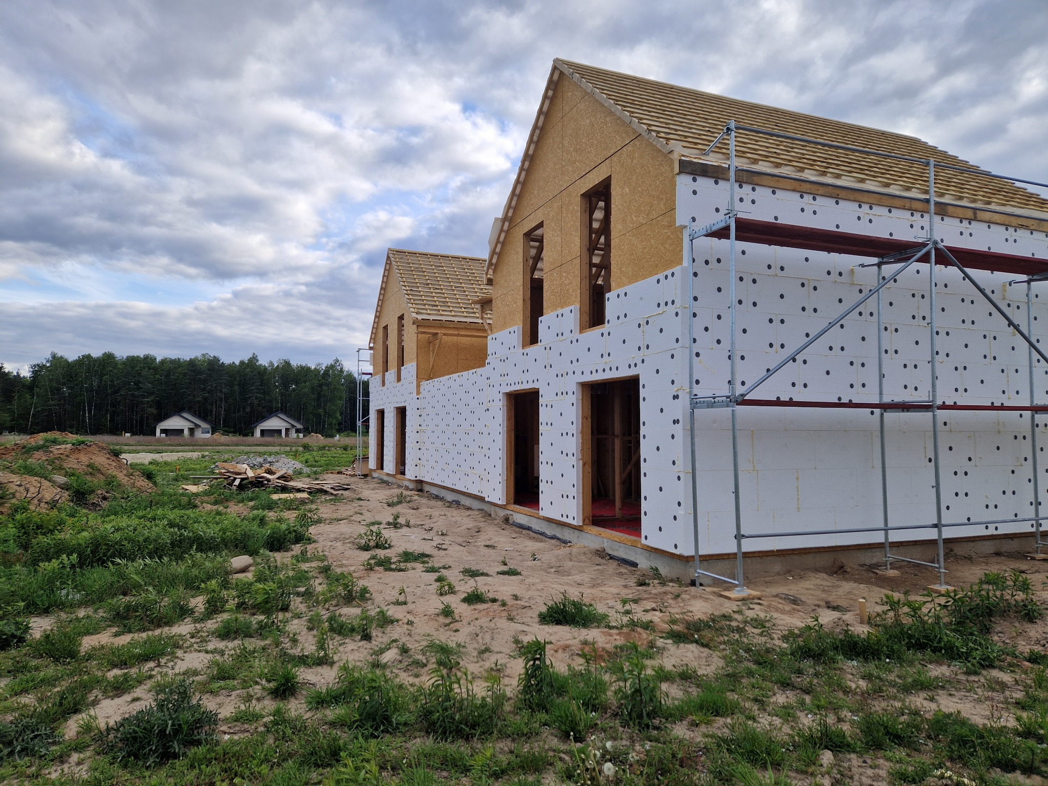 Budowa domu szkieletowego: widok na konstrukcję z płyt OSB i ocieplenia, rusztowanie przy ścianie, w tle zieleń i dwa ukończone budynki.