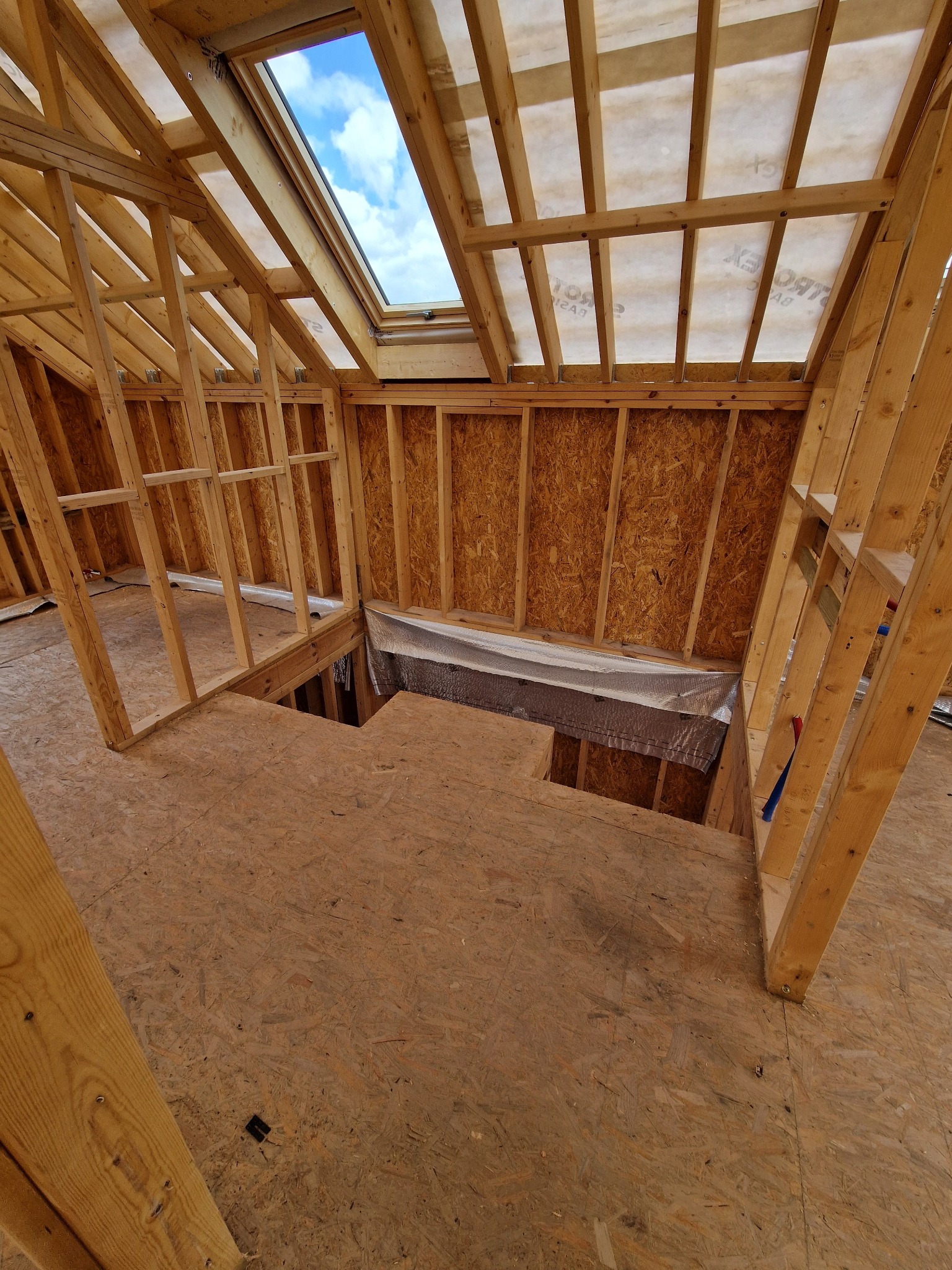 Wnętrze domu w trakcie budowy szkieletowej. Widoczna drewniana konstrukcja ścian, stropu i dachu z oknem dachowym. Elementy izolacji. Surowy stan wykończenia.