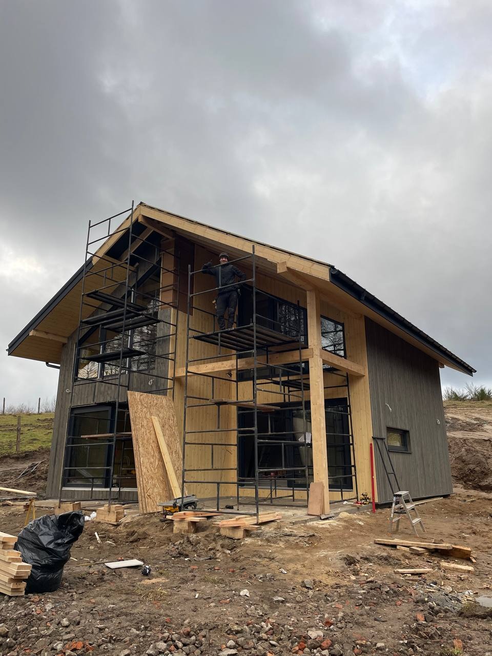 Budowa domu szkieletowego w trakcie: elewacja częściowo ukończona, widoczne rusztowania, pracownik na wysokości, drewniane elementy konstrukcyjne.
