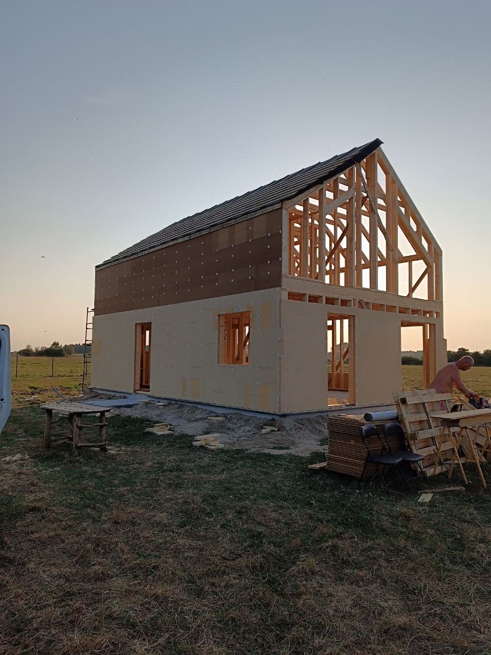 Wznoszenie domu szkieletowego: widoczna konstrukcja drewniana, częściowo pokryta płytami, z fragmentem dachu. Prace w toku, w tle łąka i zachodzące słońce.