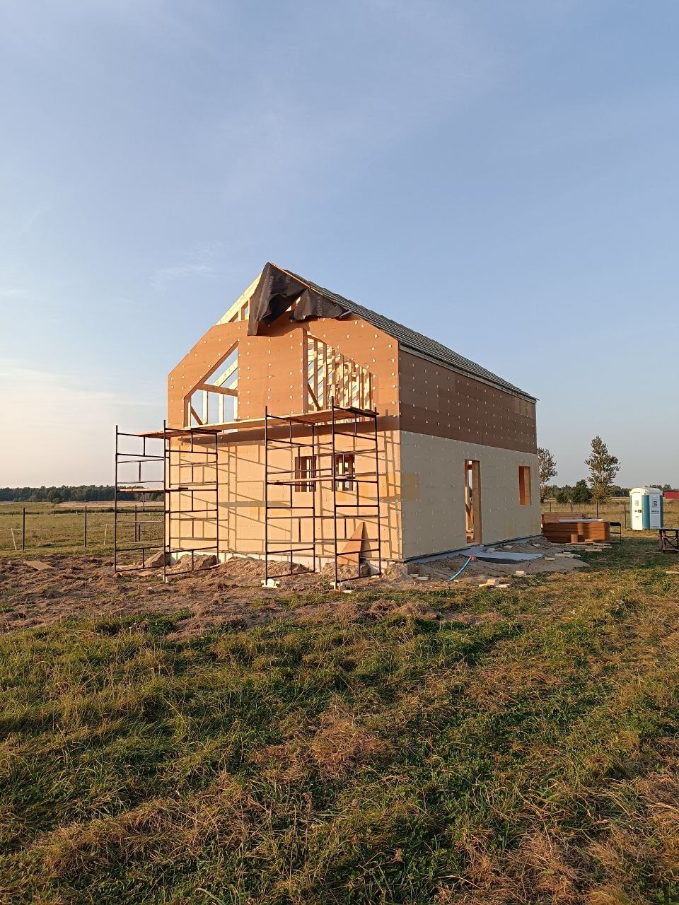 Surowy stan domu szkieletowego z widocznym drewnianym szkieletem, częściowo obity płytami. Na pierwszym planie rusztowanie, w tle zielony krajobraz.