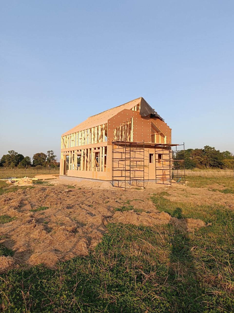 Wznoszona konstrukcja domu szkieletowego w otoczeniu zieleni. Widoczne rusztowania i elementy konstrukcyjne dachu. Częściowo pokryty dach. Słoneczny dzień.