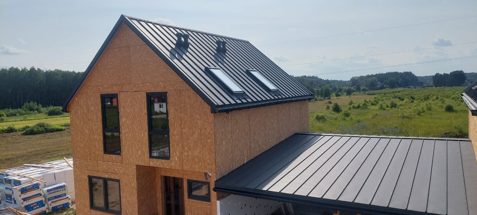 Surowy dom szkieletowy z czarnym dachem blaszanym i oknami dachowymi, otoczony zielenią. Widoczne materiały budowlane i elementy konstrukcyjne.
