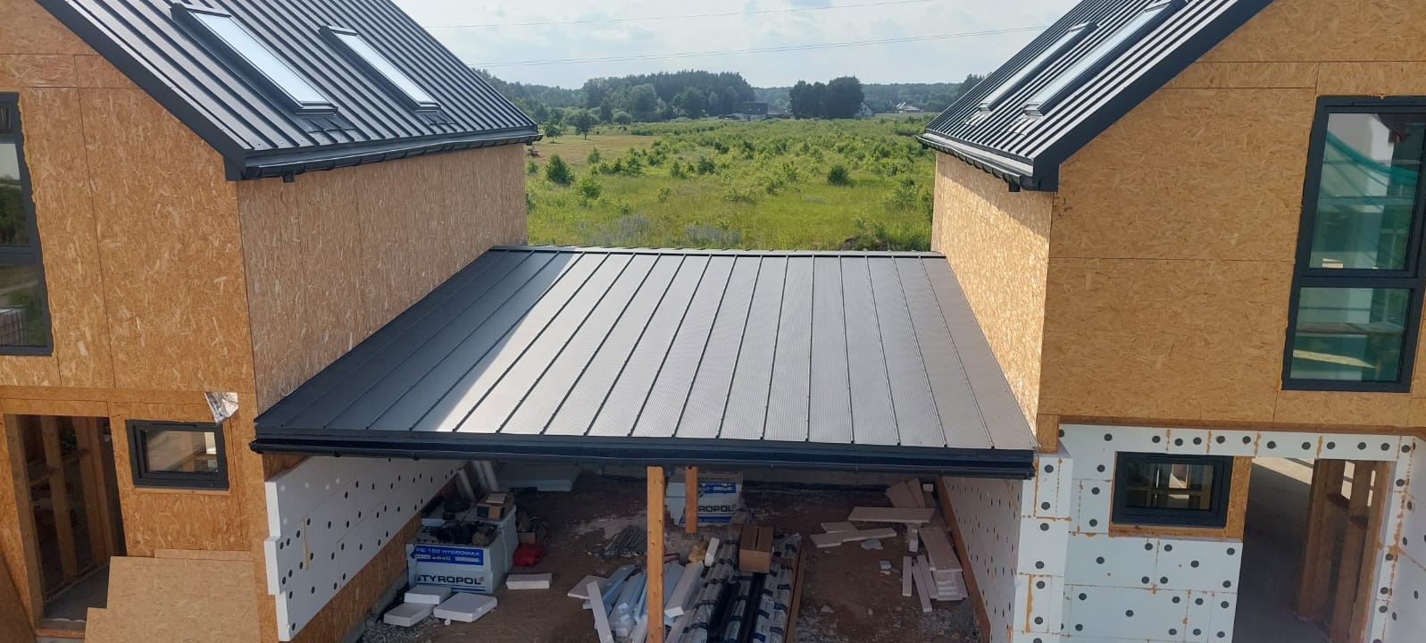 Dom szkieletowy w trakcie budowy: widok z góry na łącznik z dachem z blachy na rąbek stojący i ścianami z płyt OSB oraz oknami.