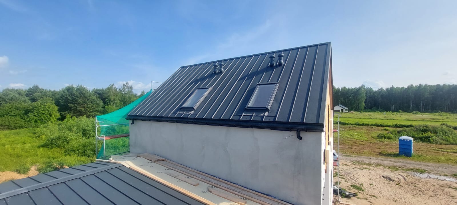 Dom w budowie z ciemnym, metalowym dachem z dwoma oknami dachowymi i wentylacją. Widoczne rusztowania i elementy konstrukcyjne.