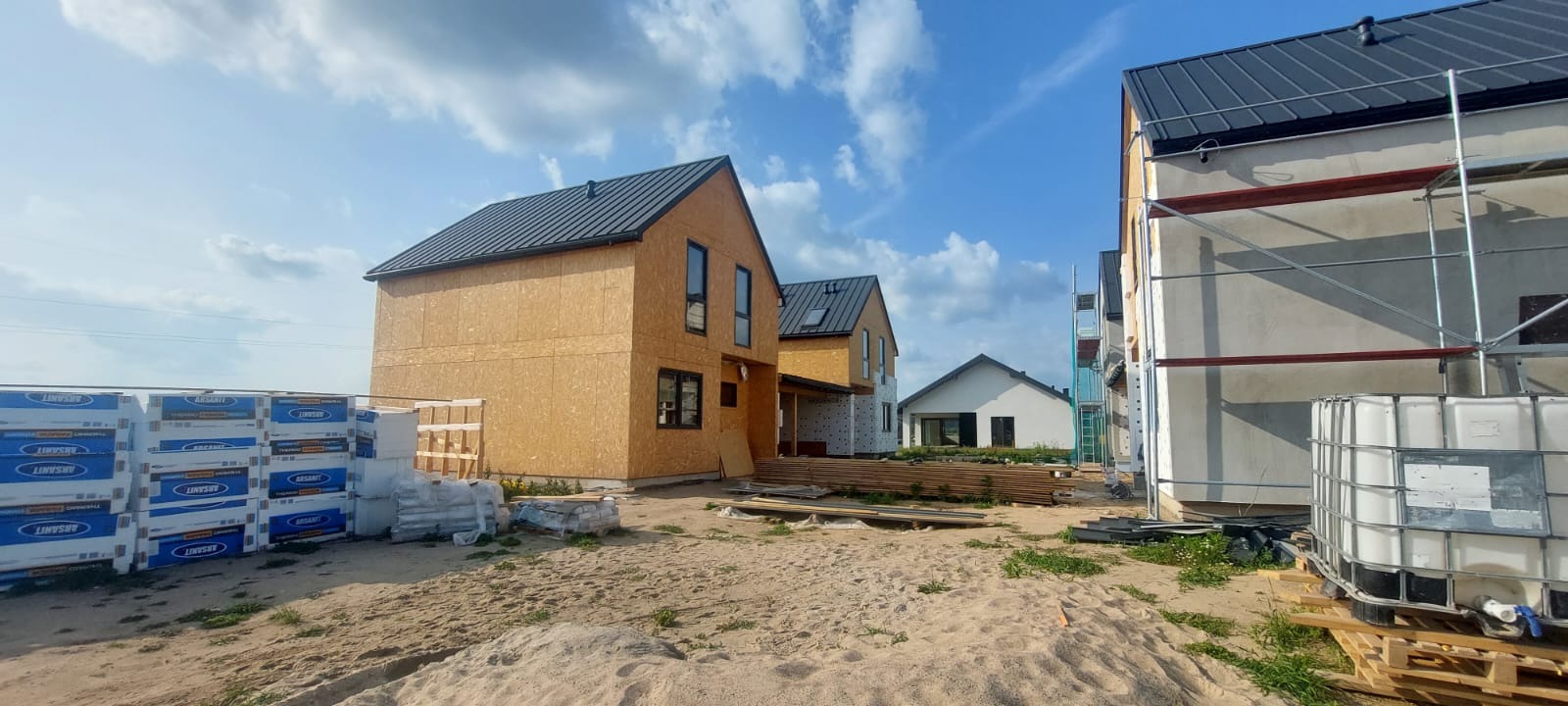 Domy szkieletowe w trakcie budowy z widocznymi płytami OSB na elewacji i czarnym dachem. Na pierwszym planie materiały budowlane i piasek.