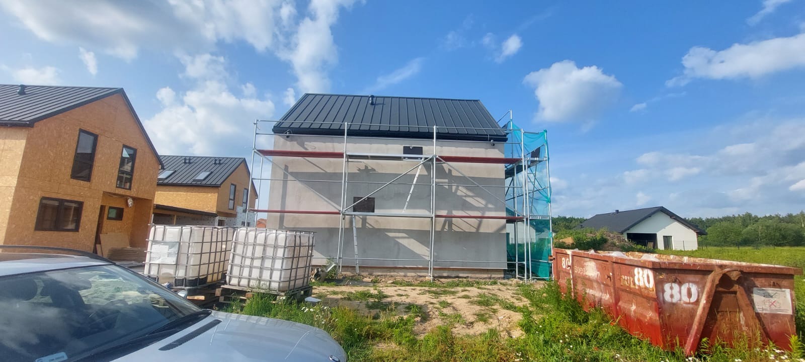 Budowa domu z szarym dachem w trakcie prac wykończeniowych, widoczne rusztowania i kontenery na gruz. W tle domy w stanie surowym i błękitne niebo.