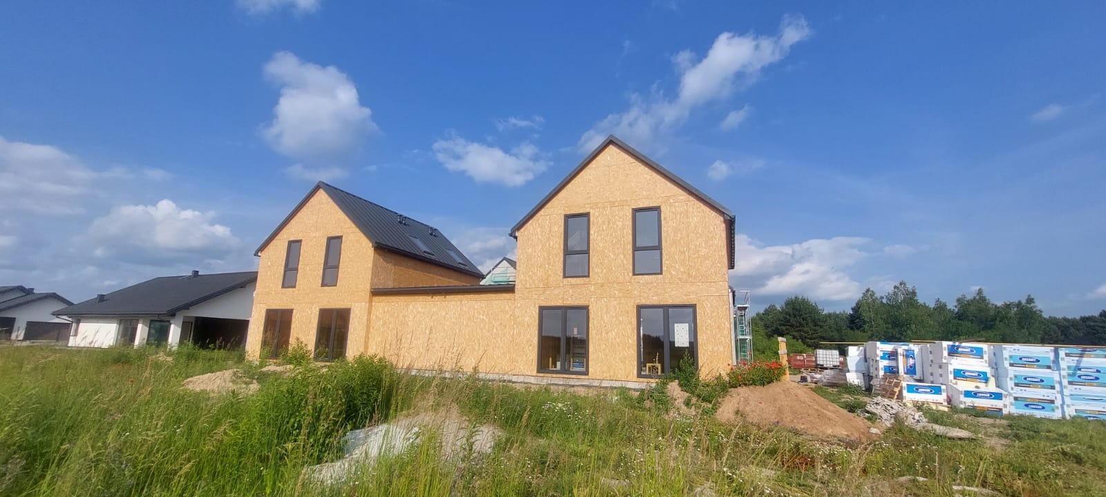 Dwa domy szkieletowe w stanie surowym zamkniętym, z czarnym dachem i oknami, na tle błękitnego nieba z chmurami. Na pierwszym planie trawa i materiały budowlane.