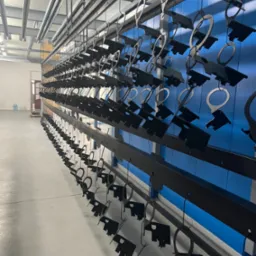 Czarne metalowe elementy zawieszone na hakach w hali, przygotowane do malowania proszkowego. Widoczna niebieska ściana w tle i metalowa konstrukcja sufitu.