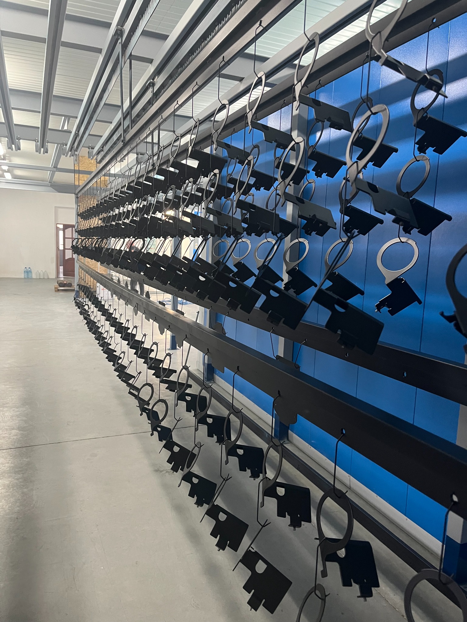 Czarne metalowe elementy zawieszone na hakach w hali, przygotowane do malowania proszkowego. Widoczna niebieska ściana w tle i metalowa konstrukcja sufitu.