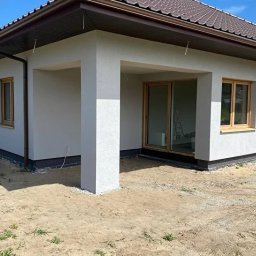 Elewarm - Jasny dom z brązowym dachem i oknami w drewnianych ramach. Widoczna elewacja po tynkowaniu i kolumna podtrzymująca zadaszenie.