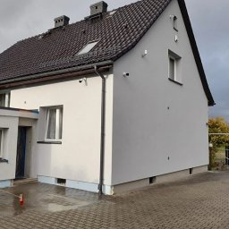 Elewarm - Dom po tynkowaniu elewacji, pracownik czyści podjazd. Widoczna świeżo otynkowana ściana budynku z oknami i czarnym dachem. W tle pochmurne niebo i drzewa.