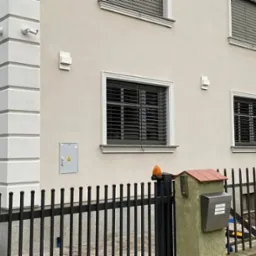 Jasna elewacja domu z ozdobnymi detalami okiennymi i narożnymi. Widoczne lampy zewnętrzne, kamery monitoringu i czarne, metalowe ogrodzenie.