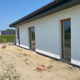 Elewarm - Jasna elewacja domu jednorodzinnego z brązowymi ramami okiennymi, widoczna z boku, z fragmentem niezagospodarowanego terenu przed budynkiem.