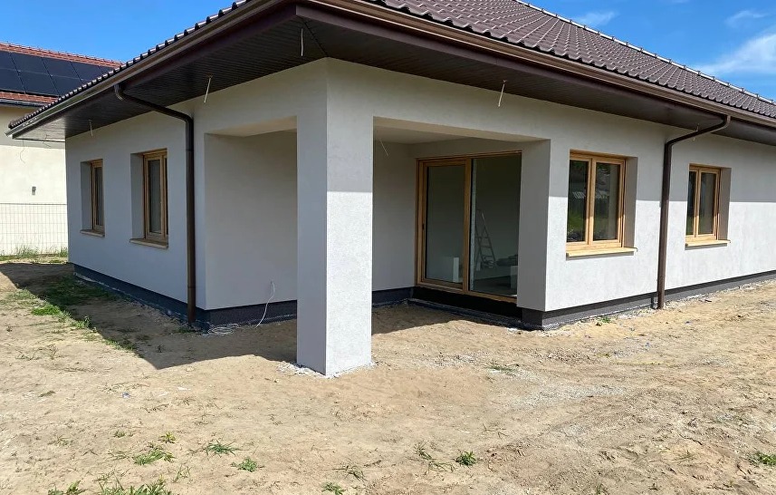 Jasny dom z brązowym dachem i oknami w drewnianych ramach. Widoczna elewacja po tynkowaniu i kolumna podtrzymująca zadaszenie.