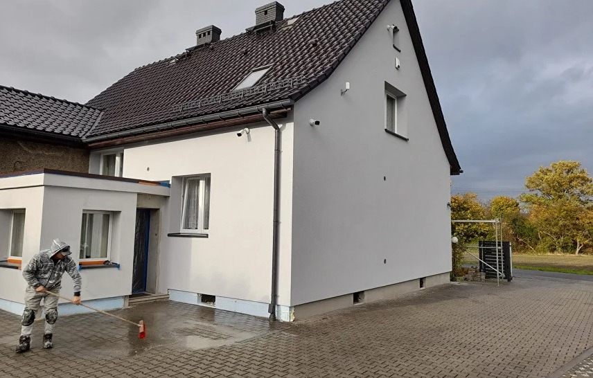Dom po tynkowaniu elewacji, pracownik czyści podjazd. Widoczna świeżo otynkowana ściana budynku z oknami i czarnym dachem. W tle pochmurne niebo i drzewa.