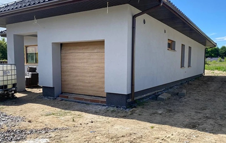 Jasna elewacja domu z drewnianą bramą garażową, widoczne detale wykończenia i elementy otoczenia. Świeżo pomalowana elewacja.