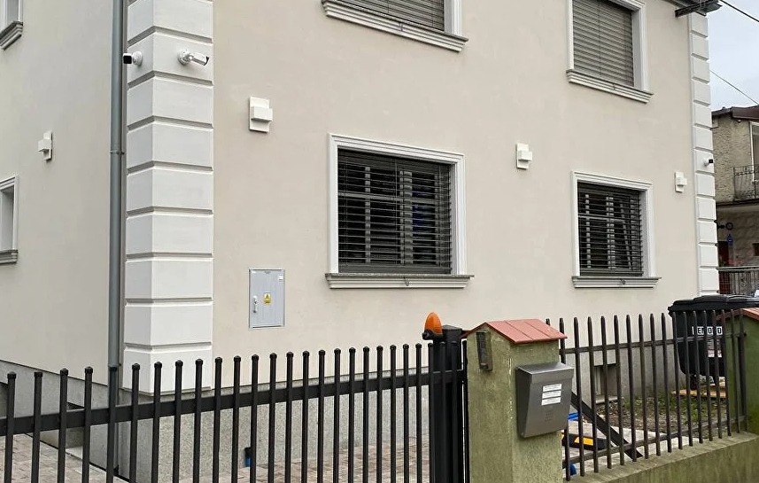 Jasna elewacja domu z ozdobnymi detalami okiennymi i narożnymi. Widoczne lampy zewnętrzne, kamery monitoringu i czarne, metalowe ogrodzenie.