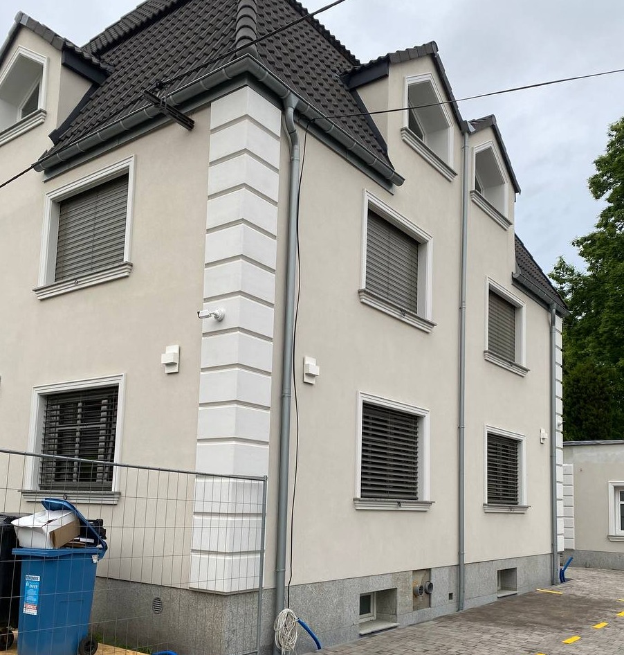 Elewacja budynku z dekoracyjnymi detalami architektonicznymi, widoczna instalacja odgromowa i fragment ogrodzenia budowlanego. Jasna kolorystyka elewacji.