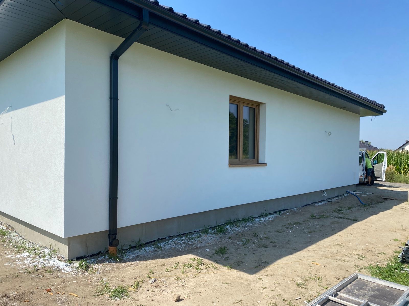 Biała elewacja domu jednorodzinnego z brązowymi oknami i czarną rynną w słoneczny dzień. Widoczne ślady po pracach elewacyjnych i samochód dostawczy.