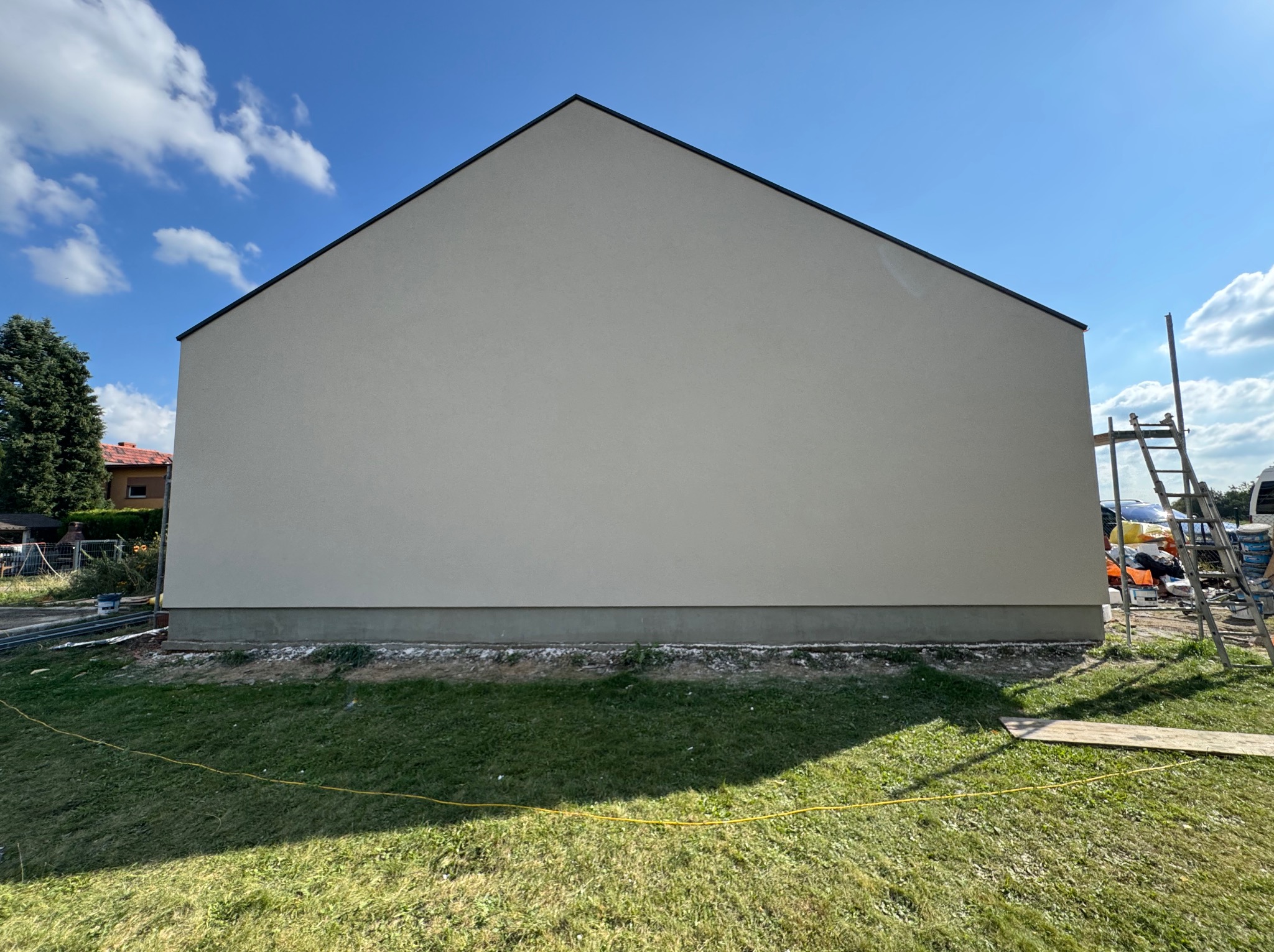 Ściana domu po tynkowaniu na gładko, widoczne rusztowanie z boku, trawa i niebieskie niebo. Minimalistyczny design elewacji.