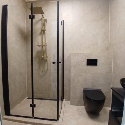 Usługi Ogólnobudowlane PERFECT BUD Aneta Zwolińska - Nowoczesna łazienka z kabiną prysznicową typu walk-in z czarnymi profilami, złotą armaturą i podwieszaną toaletą w kolorze czarnym. Jasne płytki.