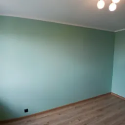 Ściana w odcieniach zieleni po malowaniu, z widocznymi listwami przypodłogowymi i sufitowymi. Na suficie widoczna lampa z kilkoma żarówkami.