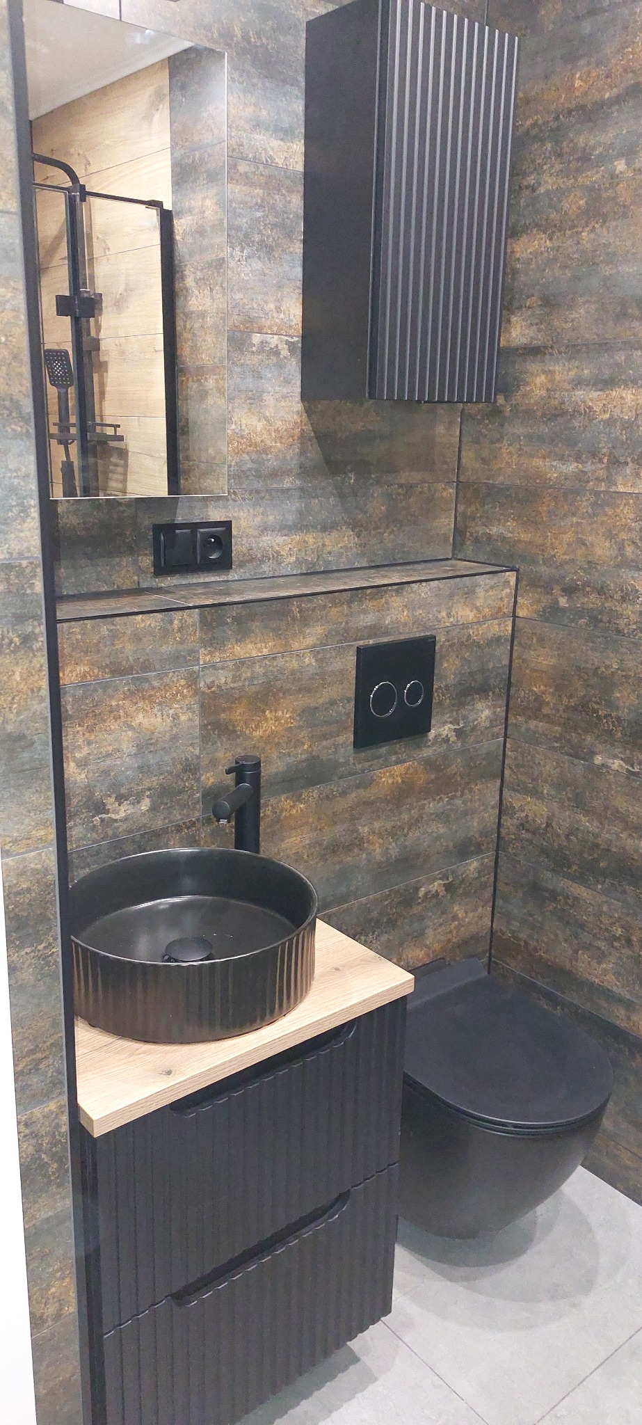 Stylowa łazienka z umywalką nablatową, toaletą w kolorze grafitu i płytkami imitującymi drewno. Widoczne detale armatury i szafka z frezowanym frontem.