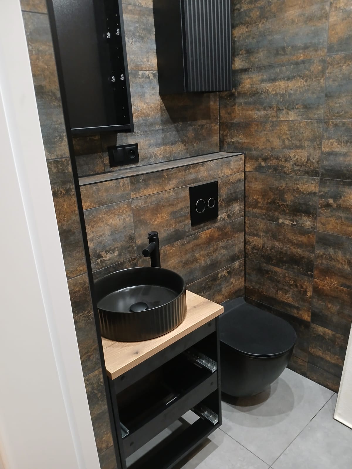 Elegancka łazienka z czarną umywalką na drewnianym blacie i wiszącą toaletą, brązowe płytki imitujące rdzę, czarne szafki, szara podłoga.