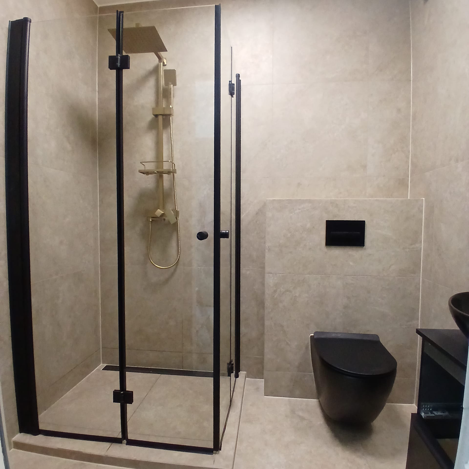 Nowoczesna łazienka z kabiną prysznicową typu walk-in z czarnymi profilami, złotą armaturą i podwieszaną toaletą w kolorze czarnym. Jasne płytki.