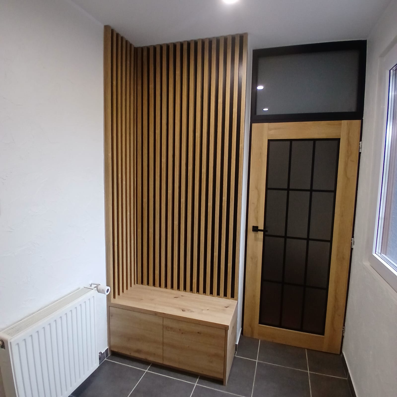 Nowoczesna aranżacja przedpokoju: drewniane lamele na ścianie, szafka z siedziskiem i drzwi z czarnymi szprosami. Szare płytki podłogowe i biały grzejnik.
