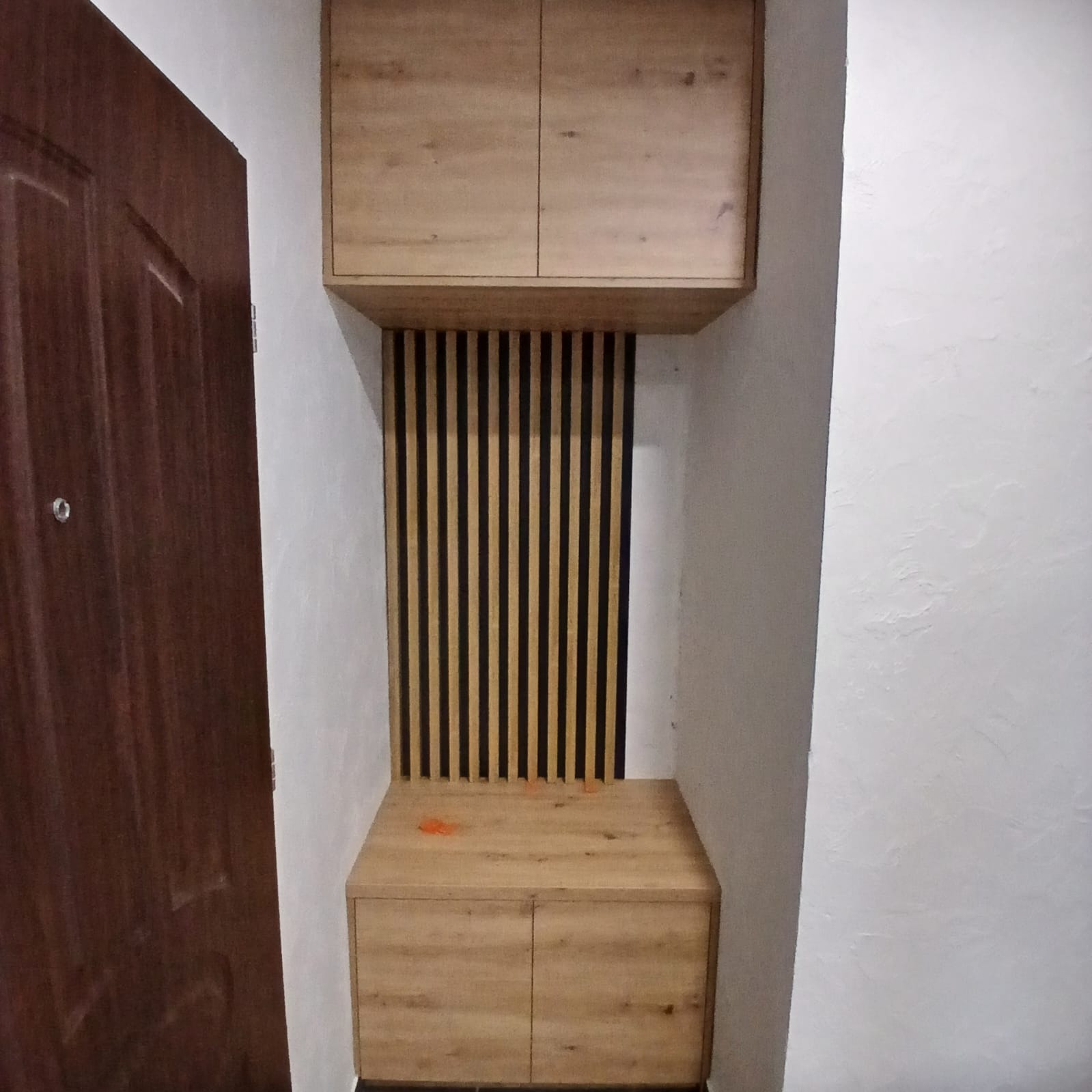 Drewniana zabudowa przedpokoju z lamelami i szafkami w odcieniach beżu, widoczna obok brązowych drzwi wejściowych na tle białej ściany.