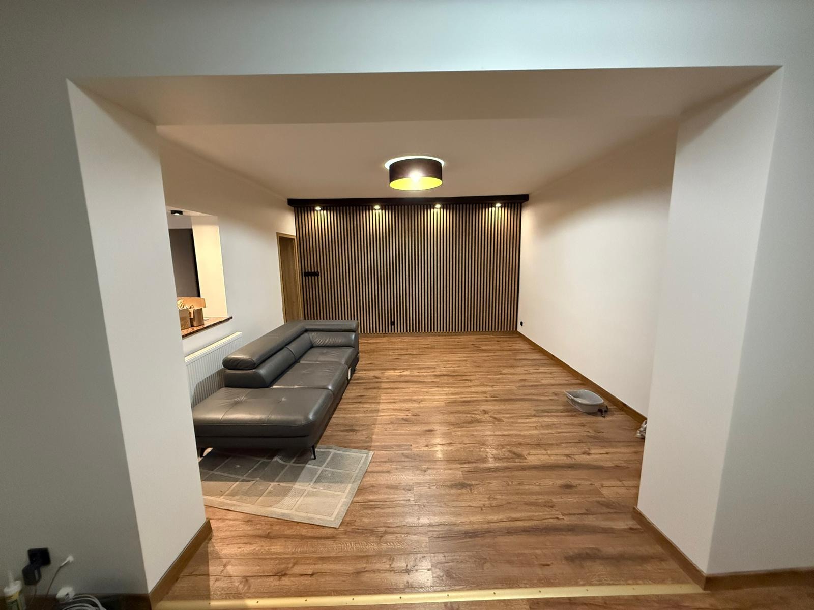 Salon po remoncie: drewniana podłoga, szara sofa, ściana z drewnianych paneli i oświetlenie. Widok z korytarza.