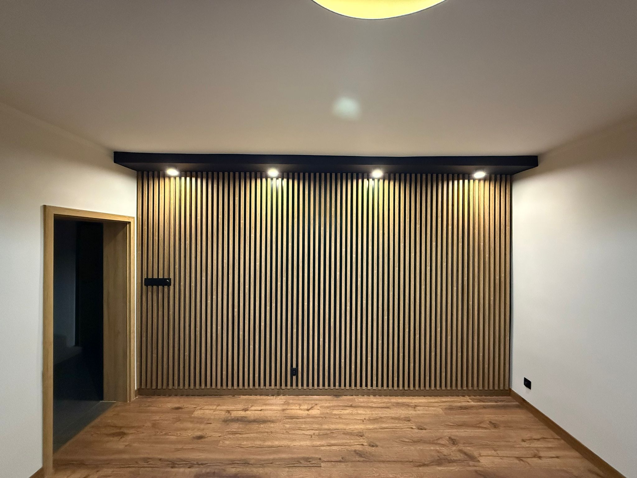 Ściana z drewnianych lameli podświetlonych punktowo, z widoczną podłogą z paneli i fragmentem drzwi. Nowoczesne wykończenie wnętrza z akcentem na naturalne materiały.