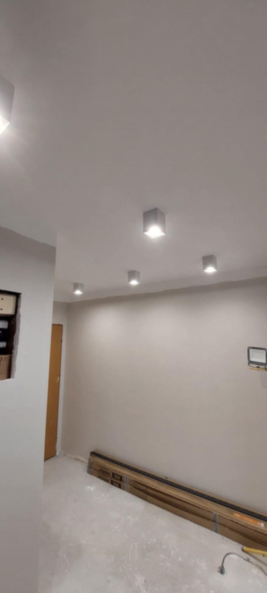 Jasne wnętrze po remoncie z czterema kwadratowymi lampami sufitowymi. Na podłodze panele podłogowe w opakowaniach. Ściany pomalowane na jasny odcień beżu.